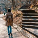 woman in brown coat and blue denim jeans walking on wooden bridge during daytime