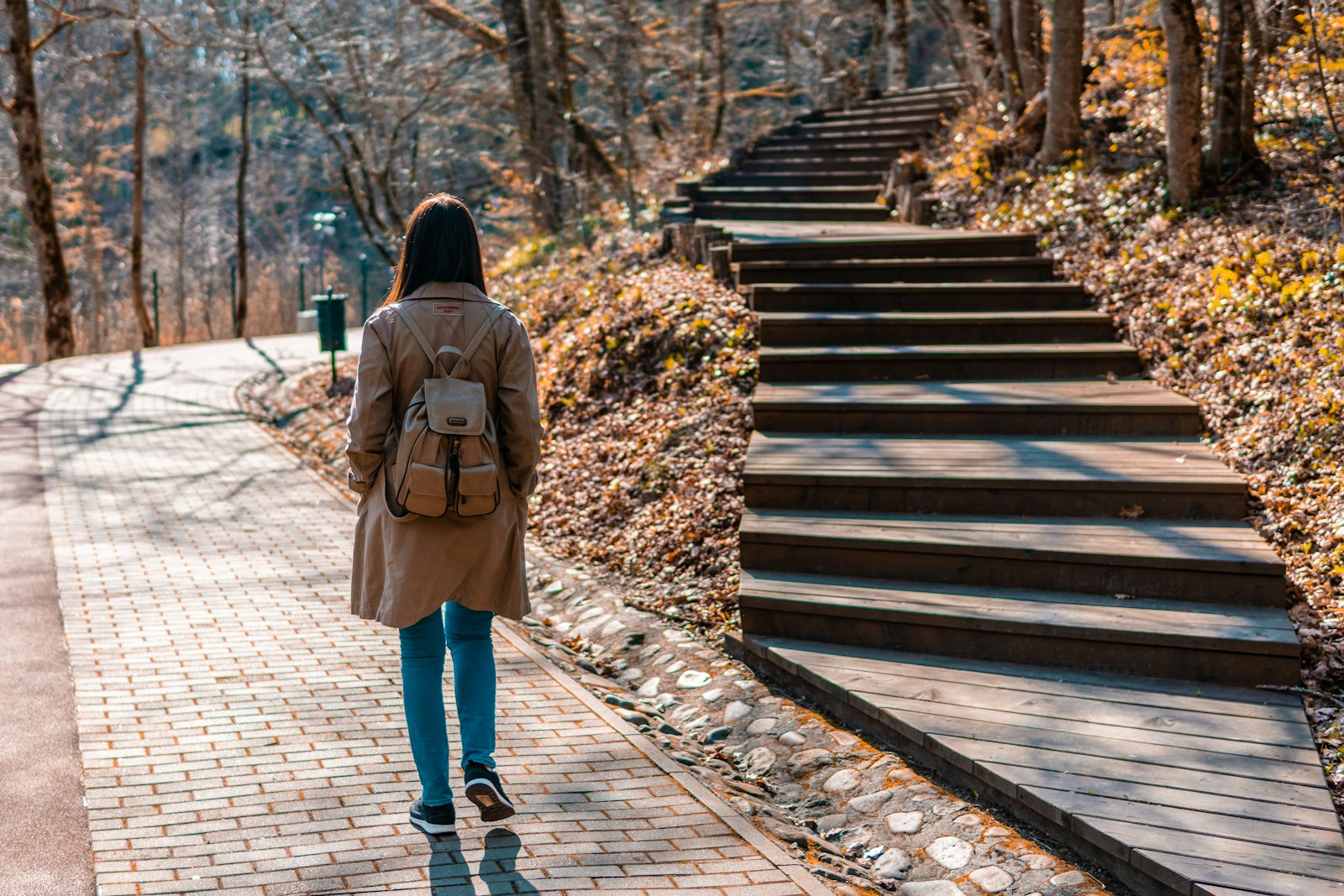 woman in brown coat and blue denim jeans walking on wooden bridge during daytime