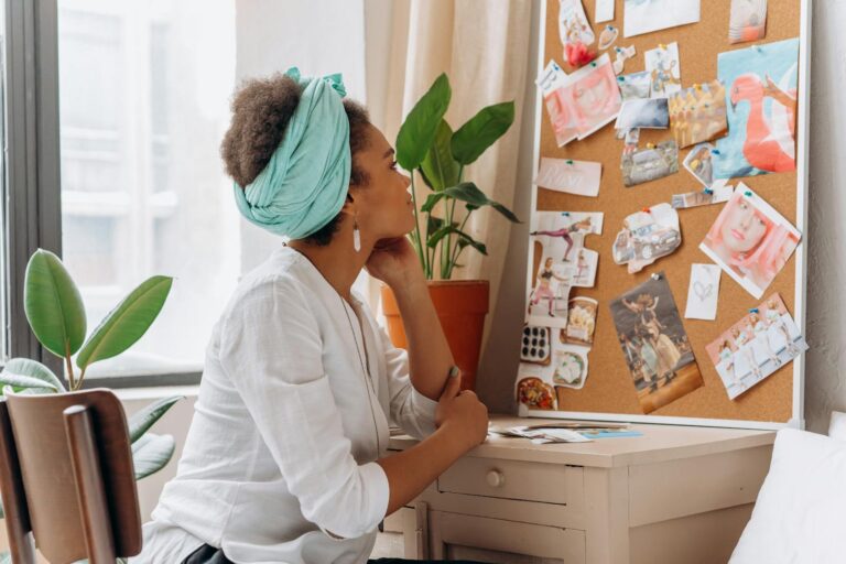 African American woman reflecting on a vision board by a window with plants.