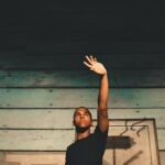 A young man stands indoors against a wooden wall, raising his hand, creating a dramatic effect.