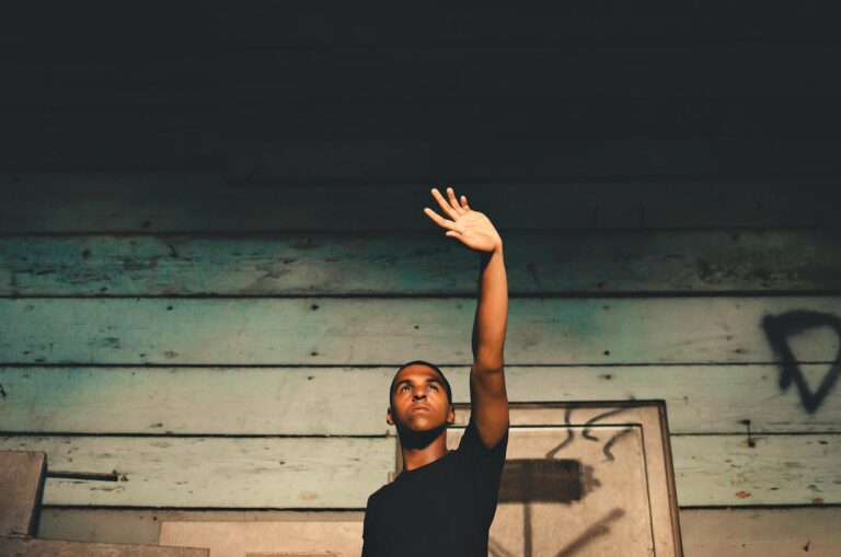 A young man stands indoors against a wooden wall, raising his hand, creating a dramatic effect.